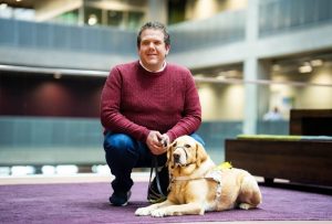 Robbie, a white male with short curly hair, is crouched down in a general office environment. Robbie is wearing a maroon jumper, blue jeans and trainers. In front of Robbie is his guide dog, lying down in harness looking at the camera.