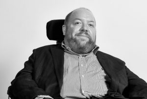 Ross a balding chap in his wheelchair in a blazer and shirt. THE IMAGE IS BLACK-AND-WHITE