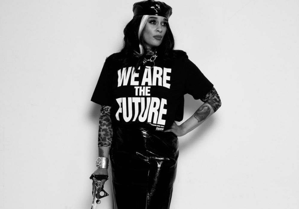 Roxy is standing confidently against a plain background. They are wearing a black beret, a black T-shirt with the bold text "WE ARE THE FUTURE" in white capital letters, and a shiny black vinyl skirt that reaches below the knees. They also have high-heeled platform boots. The Roxy's left arm has visible tattoos and is resting on their hip. Their right hand, also tattooed, is holding a long, slender walking cane with the tip resting on the ground. The overall look is edgy and fashion-forward, combining elements of punk and futuristic style.