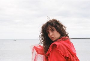 A white woman with long brown curly hair, wearing a red layered top looking over her shoulder into the camera. She is by the sea and the sky is grey behind her. She is holding some red, translucent fabric in her hand.