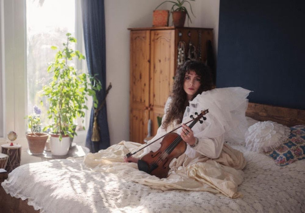 A white woman with long brown curly hair wearing a large white dress, sitting on a bed with a crochet cover. The light shines in the window behind her and there a few trees visible. She is holding a violin and there is a houseplant and wardrobe in view.