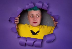 White woman with short hair with green fringe bursting through a purple background. yellow top with a large crow necklace.