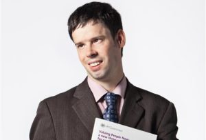 White man smiling brown hair holding a book under left arm suit brown