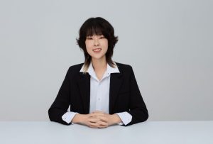 An Asian person with shoulder-length hair smiles while wearing a black blazer and white shirt, sitting at a table with hands clasped.