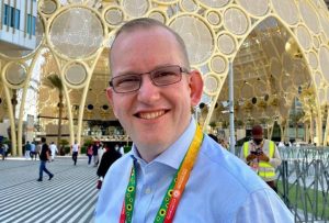 A blond haired man wearing glasses and blue shirt in front of Al Wasl dome, Dubai