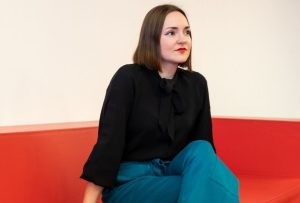 A white woman with brown bob cut hair is sat on a red sofa. She is wearing a black shirt, turqouise culottes, black high heels and red lipstick.