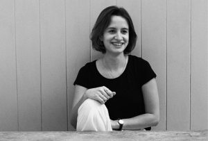 A young woman with dark hair in a bob is sitting on a bench and smiling. She is wearing a black t-shirt and white trousers.