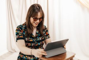 A young woman with shoulder length brown hair and dark glasses is looking down at a laptop.