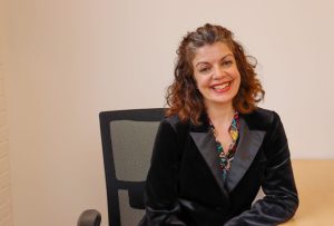 A smiling 50 year old woman with salt and pepper shoulder length hair wearing a black velvet jacket sitting at a desk