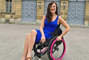 Georgina is a white, brunette female who uses a manual wheelchair. She is smiling and wearing a bright blue fitted dress. Her manual wheelchair has a pop of colour with baby pink pushrim covers to help her self-propel.