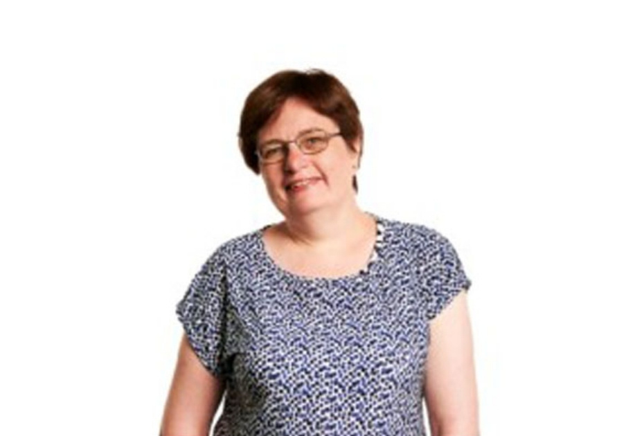 A white woman with dark hair and glasses wearing a patterned top in front of a white background.