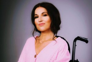 A white woman with dark brown hair sits in her wheelchair, smiling to camera.