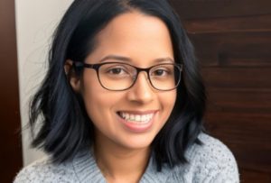 The image shows a south Asian young woman with shoulder-length black hair and glasses, wearing a light gray knitted sweater. She is smiling and looking directly at the camera. The background is a mix of a wooden panel and a light-beige coloured wall.