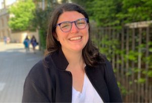 White woman with brown hair and blue glasses stands smiling in a white shirt and black blazer.