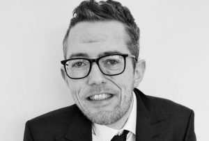 A white man in a suit, leaning against a white wall smiling to camera.