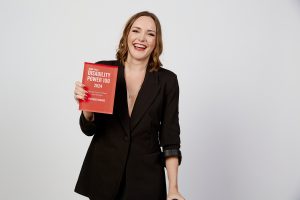 Photo of Victoria Jenkins, she is wearing a dark suit and is holding her DP100 trophy. She has dark hair and is smiling widely at the camera